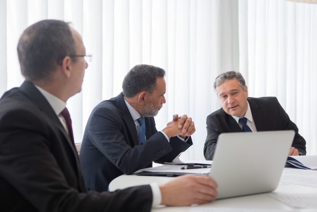 Three people in business suits talking across a table, with a laptop and printed papers.