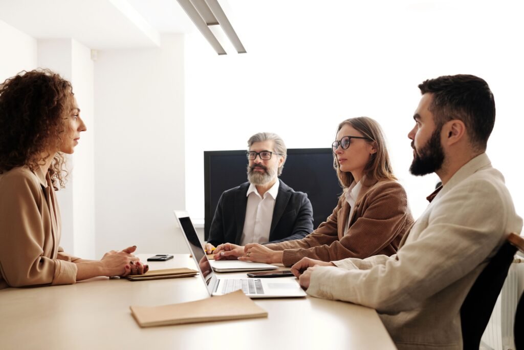 Four people around a meeting table in a bright room, talking, with a laptop open.