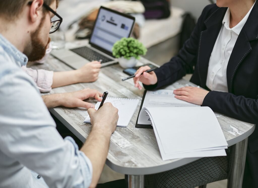 Two people at a small round table, one writing in a notebook while the other holds an open folder.