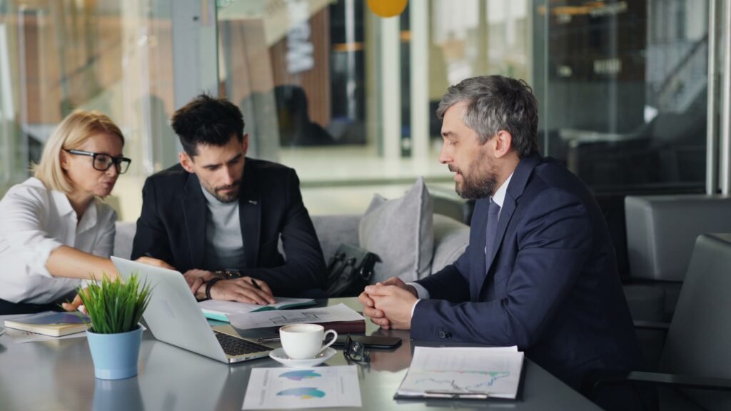 Three people at an office table, two looking at a laptop while the third listens.