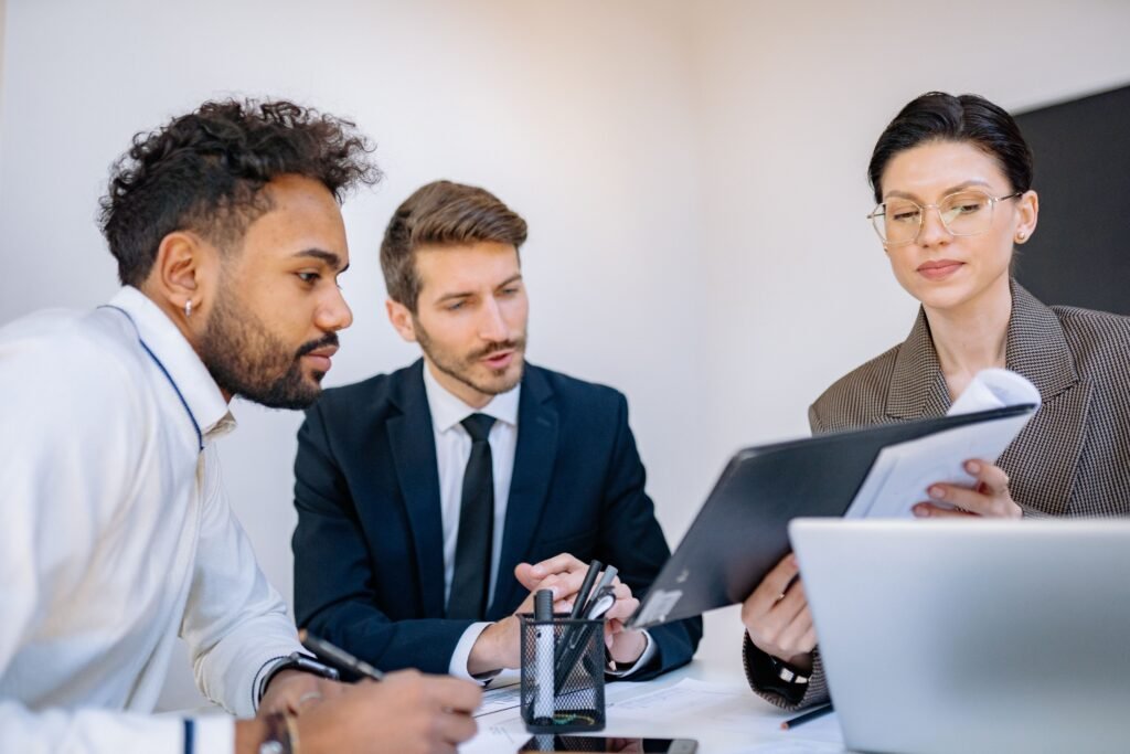 Three people looking at an open folder held up by one of them, with a laptop on the desk.