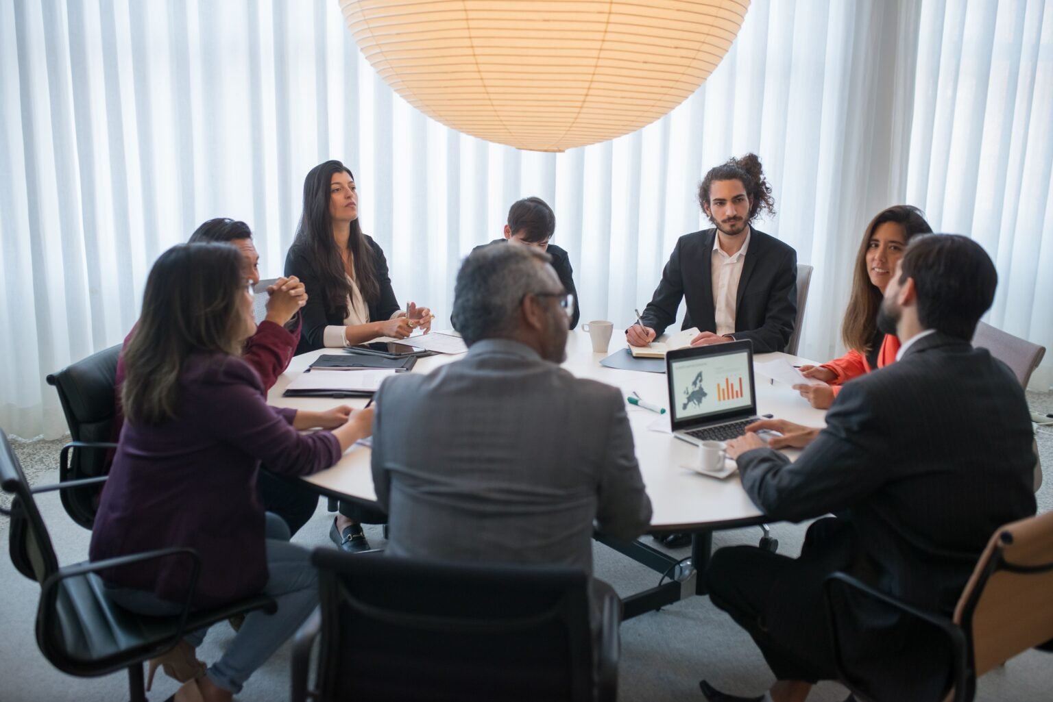 Eight people seated around a large round meeting table with laptops and notes.